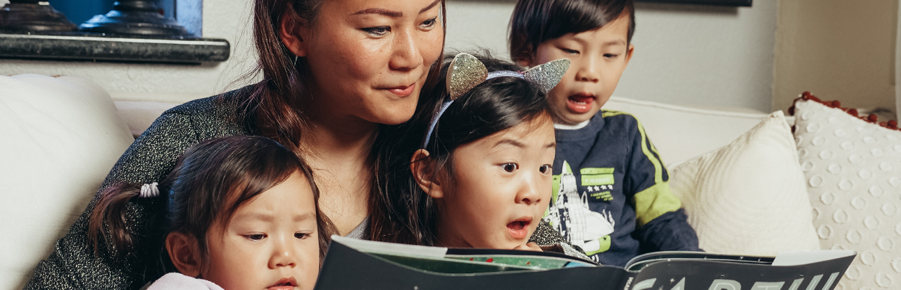 A mother and her three young children sitting on a couch in front of a laptop enjoying Story Time virtually.